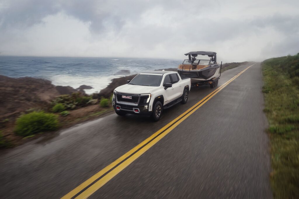 Front 3/4 view of the 2026 GMC Sierra EV AT4 in Summit White towing a boat on a seaside road.