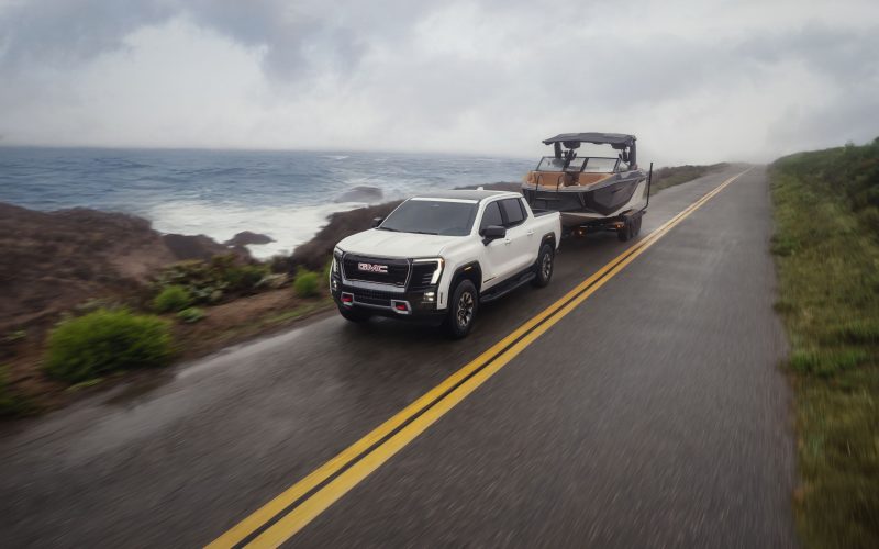 Front 3/4 view of the 2026 GMC Sierra EV AT4 in Summit White towing a boat on a seaside road.