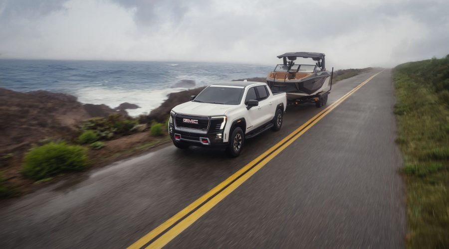 Front 3/4 view of the 2026 GMC Sierra EV AT4 in Summit White towing a boat on a seaside road.